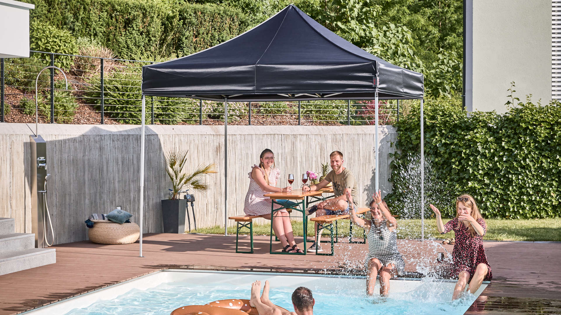 Im Garten wurde der schwarze 3x3 m Faltpavillon und die klassische Bierzeltgarnitur aufgestellt. Freunde verbringen im Garten und im Pool den Tag.