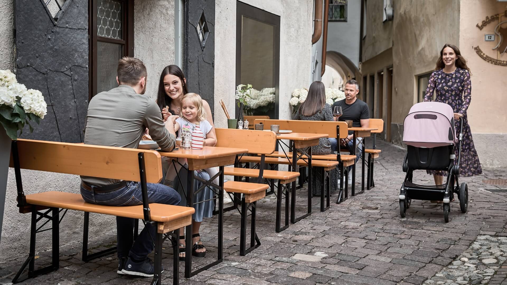 Several people sitting on small beer garden table sets in an alley near a restaurant.