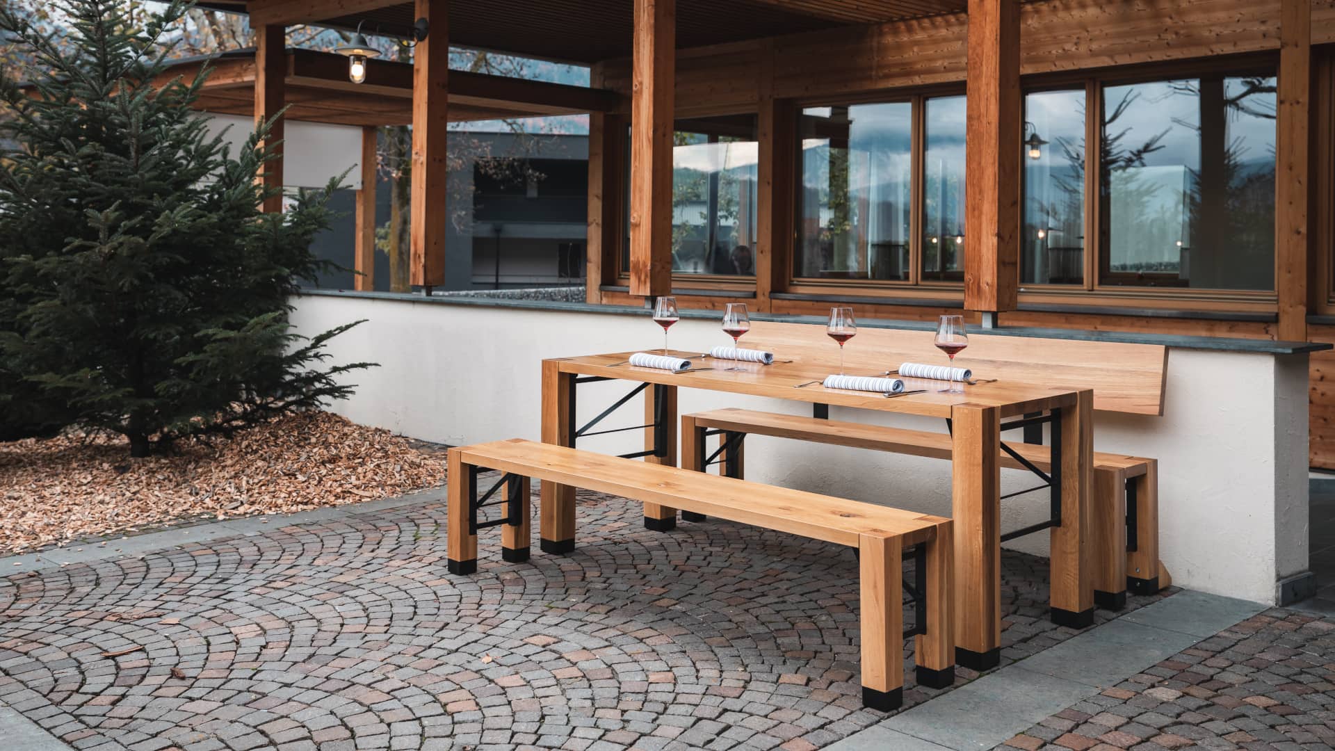 Design set Lago is placed in front of a building. One bench with backrest and one without backrest, the table is set.