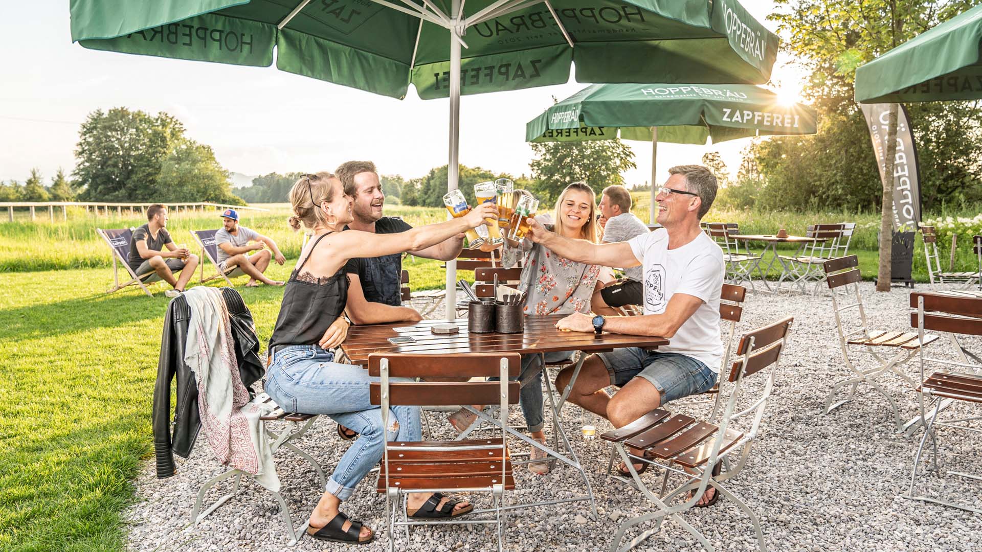 4 Personen stoßen mit ihrem Bier auf den Biergartenmöbeln auf der Terrasse eines Lokals an.