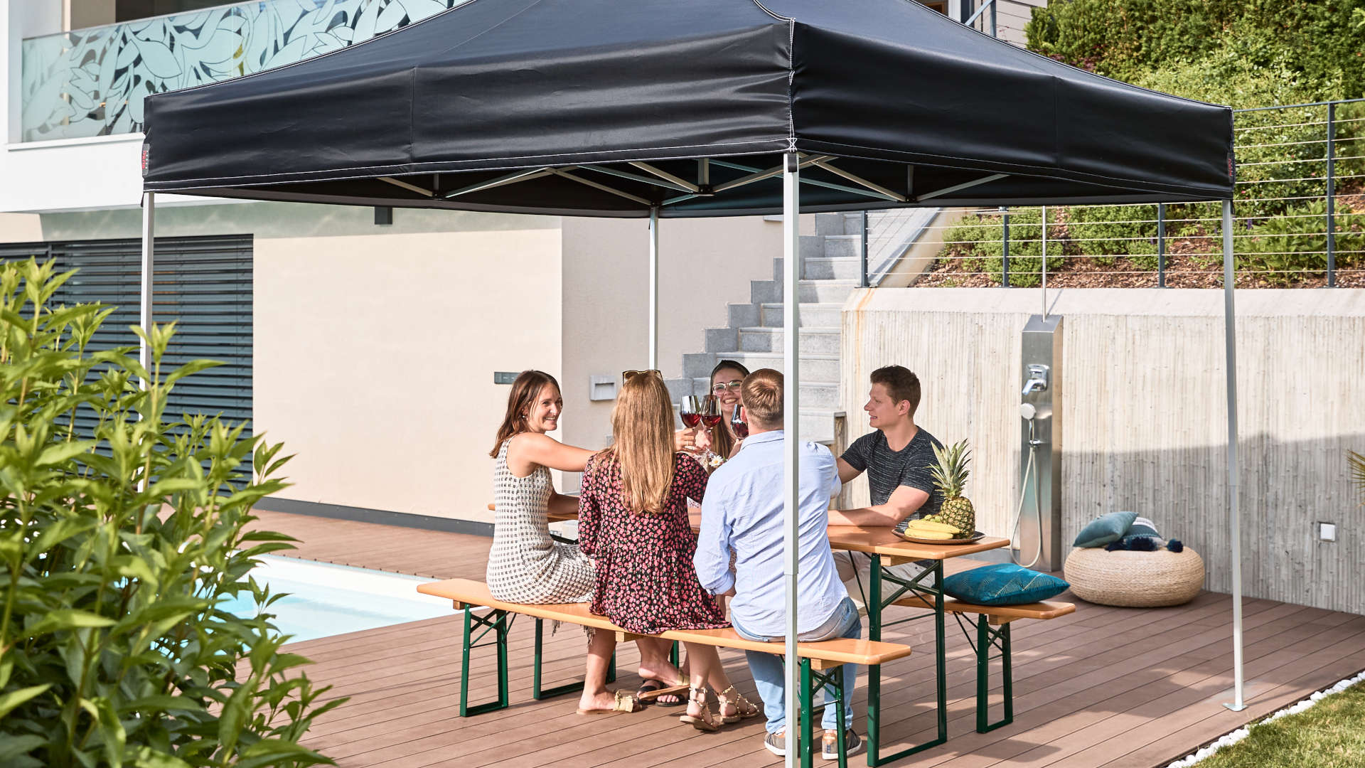 Im Garten wurde der schwarze 3x3 m Faltpavillon und die klassische Bierzeltgarnitur aufgestellt. Freunde verbringen im Garten vor dem Pool den Tag.