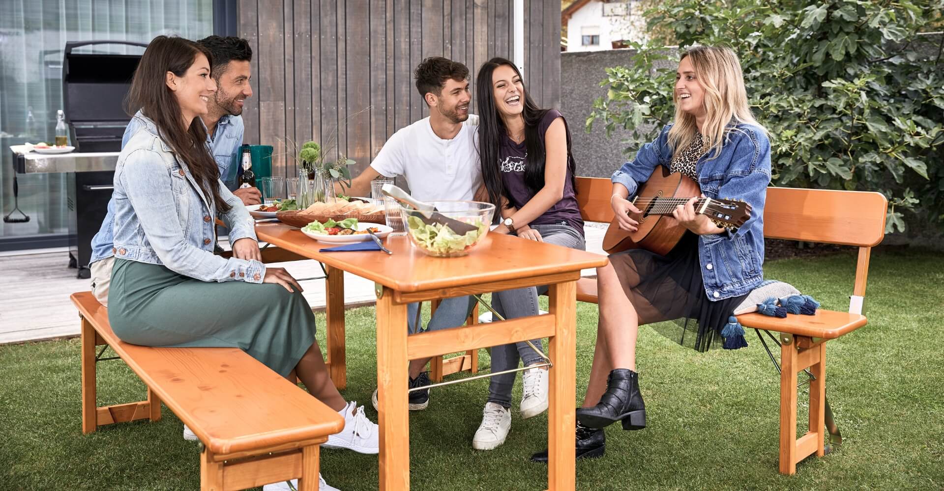 Four people enjoying the music played by a woman on a guitar sitting on the design set Rustica in the garden.