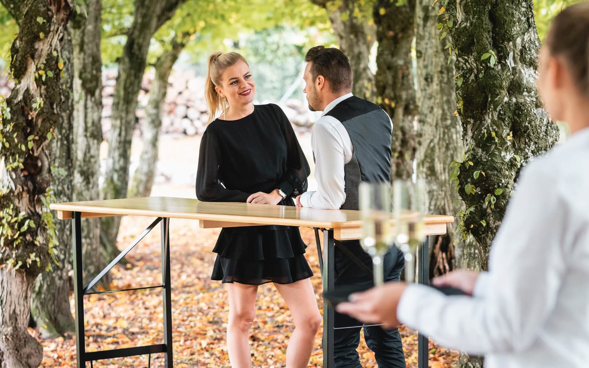 Zwei Personen stehen am Stehtisch und warten auf eine weitere Person, die mit Gläsern zu ihnen kommt. 