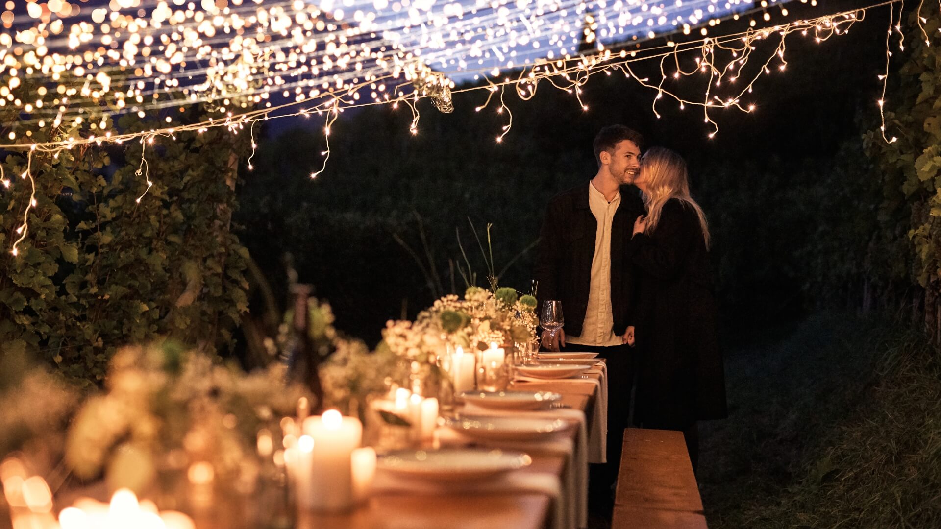 A wide beer garden table set is romantically decorated and a couple wants to spend a nice evening outside.