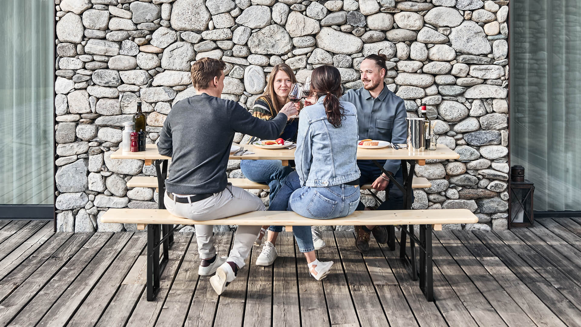 Auf einer Terrasse genießen 4 Personen Ihre Mahlzeit gemütlich auf der klassischen Bierzeltgarnitur in Natur
