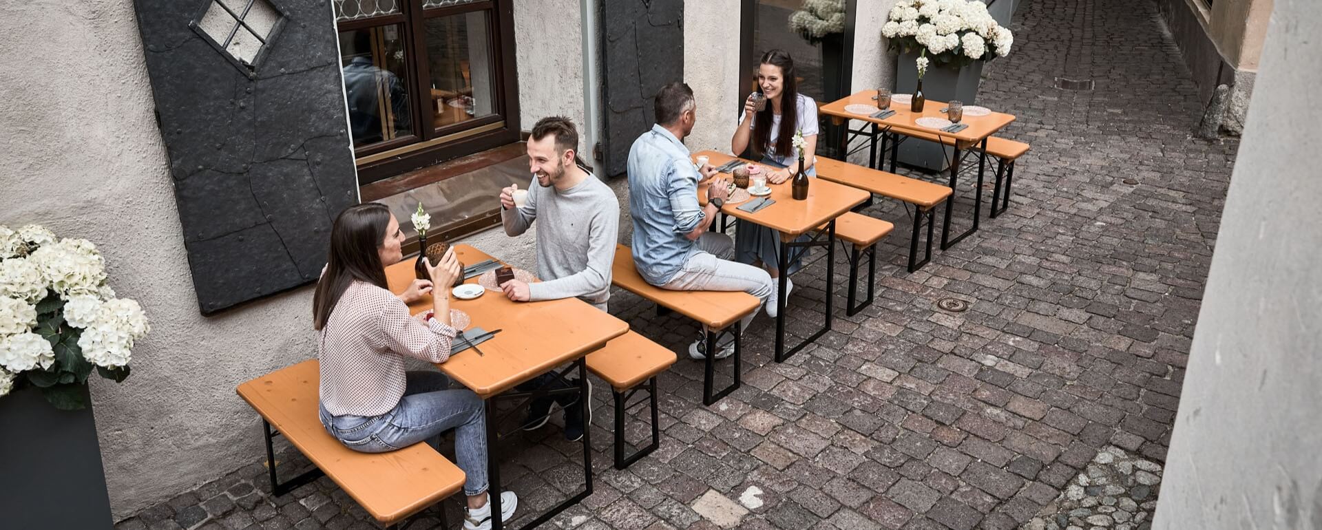 Die kleinen Bierzeltgarnituren Shorty sind im Außenbereich von einem Restaurant in einer Gasse aufgestellt und die Kunden genießen Ihre Getränke.