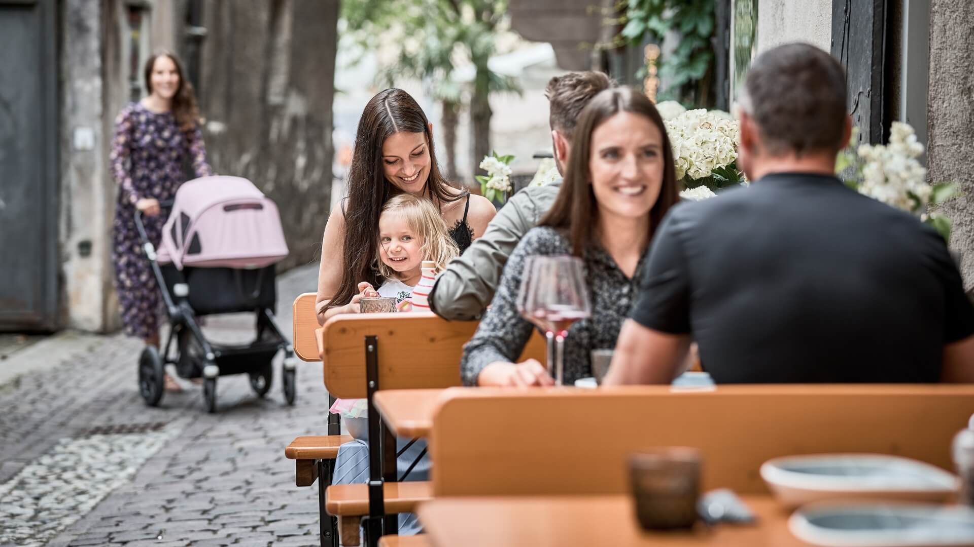 Several people are sitting on small beer garden table set in an alley near a restaurant.