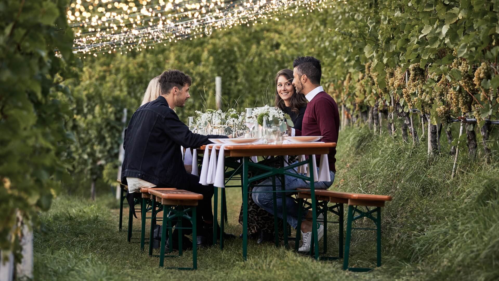 Vier Personen haben auf einer dekorierten breiten Bierzeltgarnitur Platz genommen und genießen den Tag  draußen in der Natur.