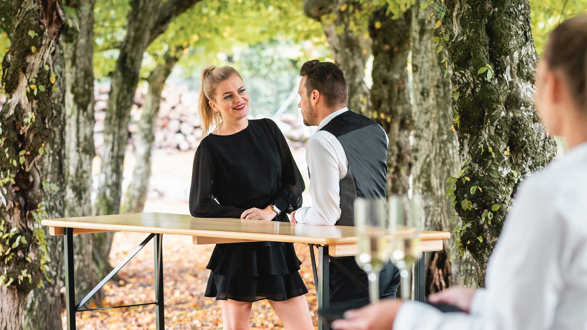 Two people are poseur at the bar table waiting for another person to join them with glasses. 