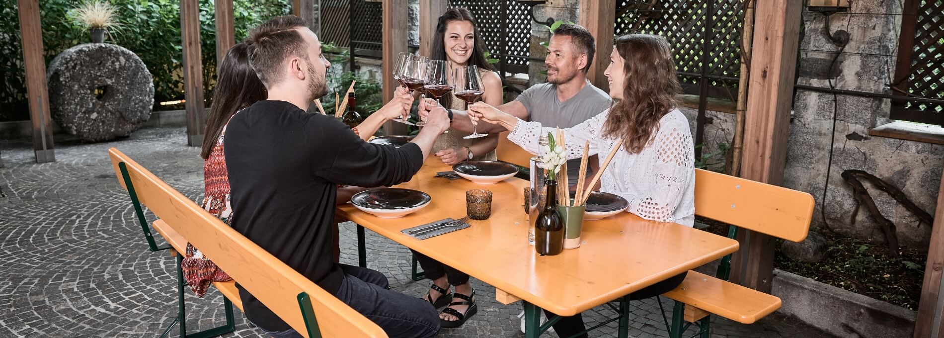 Five people are sitting in the outdoor area of a restaurant on a wide beer table set with a backrest, clinking glasses of red wine.