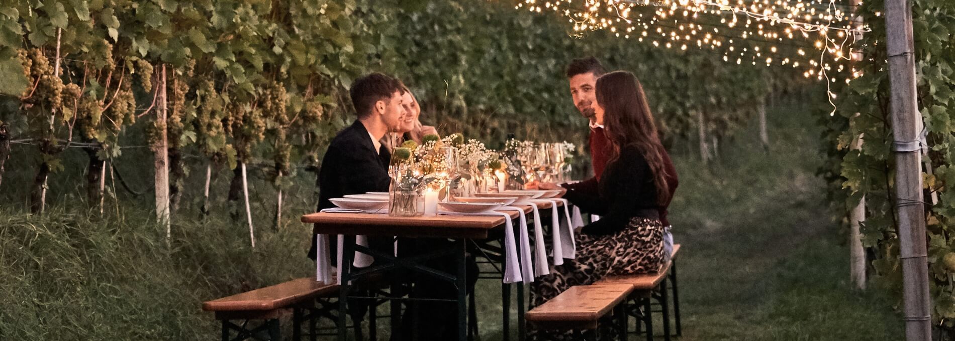 Four people sitting in the greenery between the vineyards on a beer garden table set and enjoy your meal.