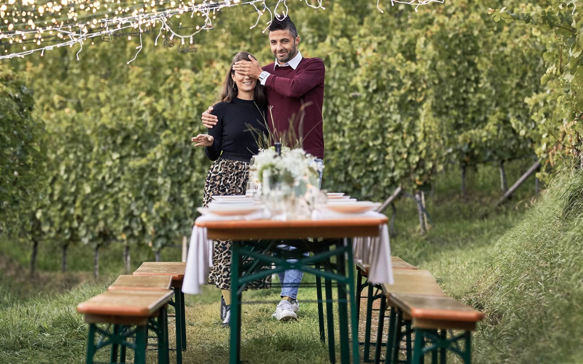A couple is walking to a decorated beer garden tabel set and the woman has the eyes closed because it has to be a surprise.