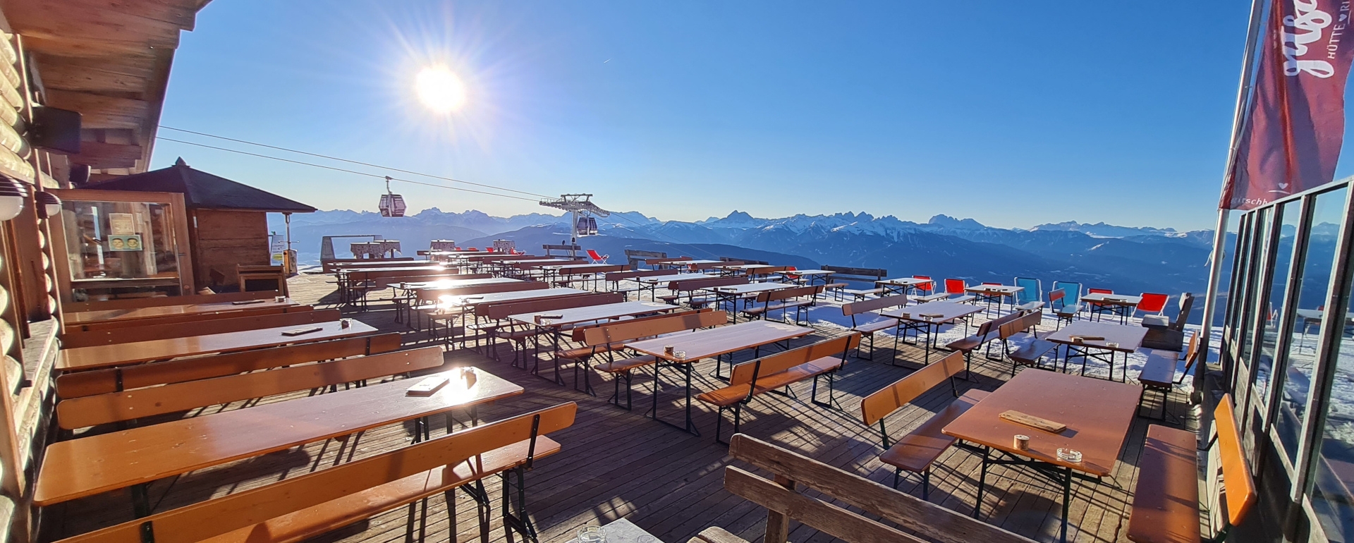 Wide beer garden table sets with backrest are placed on an alpine pasture in front of a hut in winter.