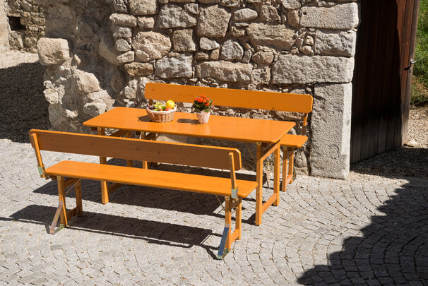 Das Bild zeigt die Designgarnitur Rustica mit Lehne in Mühlbach. Die Designgarnitur steht vor einer rustikalen Steinwand im Außenbereich und der Tisch ist mit einem Blumentopf und einem Obstkorb dekoriert.