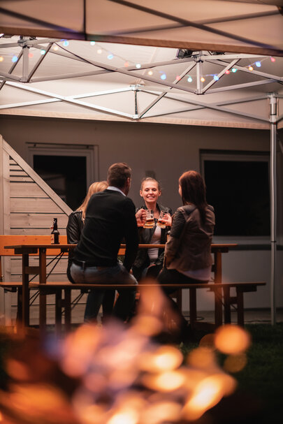 Eine gesellige Runde von vier Personen sitzt auf einer Rustica-Sitzgruppe unter einem beleuchteten wasserdichten Faltpavillon im Garten. Es ist bereits abends und dunkel. Der Faltpavillon wurde zudem mit einer Girlande aus bunt leuchtenden Glühbirnen verziehrt.