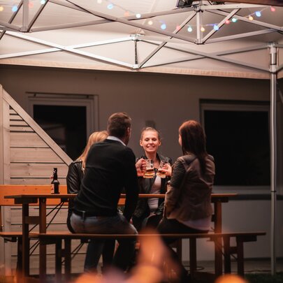 Eine gesellige Runde von vier Personen sitzt auf einer Rustica-Sitzgruppe unter einem beleuchteten wasserdichten Faltpavillon im Garten. Es ist bereits abends und dunkel. Der Faltpavillon wurde zudem mit einer Girlande aus bunt leuchtenden Glühbirnen verziehrt.