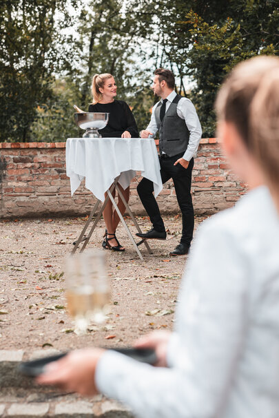 Zwei Personen werden an einem klappbaren Stehtisch, gedeckt mit einer Tischdecke, verköstigt.