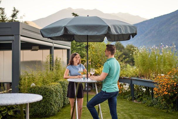 Zwei Personen genießen ihren Wein und stehen dabei an dem Stehtisch Unkaputtbar in einem Garten.