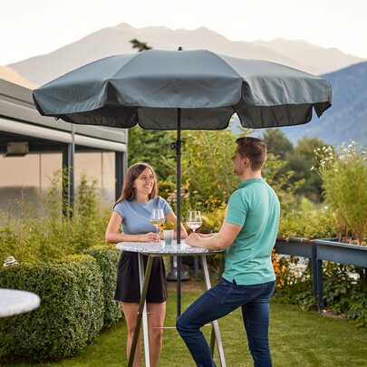 Zwei Personen genießen ihren Wein und stehen dabei an dem Stehtisch Unkaputtbar in einem Garten.