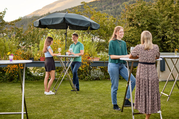 In einem Garten sind mehrere Stehtische Unkaputtbar aufgestellt und an einzelnen Tischen genießen Personen einen Aperitif und unterhalten sich.