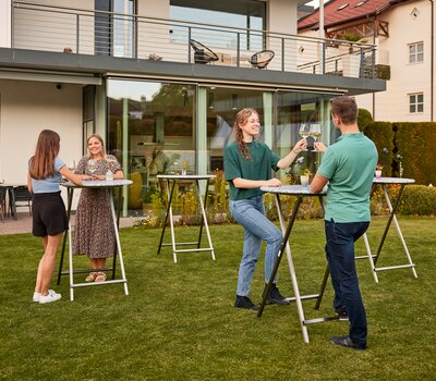 Es sind mehrere Stehtische Unkaputtbar aufgestellt und an einzelnen Tischen genießen Personen einen Aperitif und unterhalten sich.