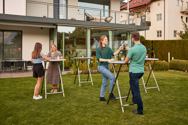 Es sind mehrere Stehtische Unkaputtbar aufgestellt und an einzelnen Tischen genießen Personen einen Aperitif und unterhalten sich.
