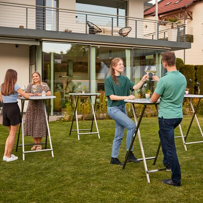 Es sind mehrere Stehtische Unkaputtbar aufgestellt und an einzelnen Tischen genießen Personen einen Aperitif und unterhalten sich.