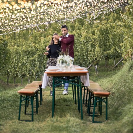 Ein Paar geht zu einer geschmückten Biergartentischgarnitur und die Frau hat die Augen geschlossen, weil es eine Überraschung sein soll.