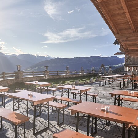 Auf einer Terrasse einer Hütte stehen kleine Bierzeltgarnituren in  der Sonne.