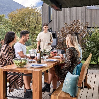 Es findet ein Grillnachmittag im Garten statt und 4 Personen sitzen bereits auf der Designgarnitur Lago und ein Mann kommt mit einem Teller auf sie zu.