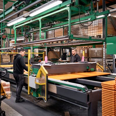 Das Bild zeigt das Innere der Produktionshalle in Schabs. Man sieht zwei Mitarbeiter, die an einer Maschine arbeiten und die Bierbänke und -tische für den nächsten Arbeitsschritt vorbereiten.