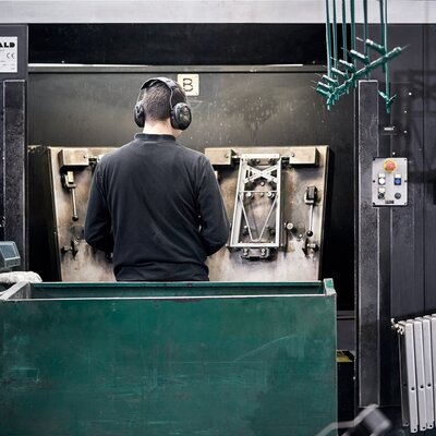 Ein Mitarbeiter ist von hinten in der Werkstatt zu sehen. Vor ihm steht eine Maschine, in der die Untergestelle für die Bierzeltgarnituren gefertigt werden.