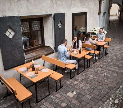 Drei Bierzeltgarnituren stehen vor einem Restaurant in einer Gasse. Die Tische sind gedeckt und an zwei von drei Tischen sitzen Leute.