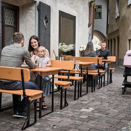 Mehrere Personen sitzen auf kleinen Bierzeltgarnituren in einer Gasse bei einem Restaurant.