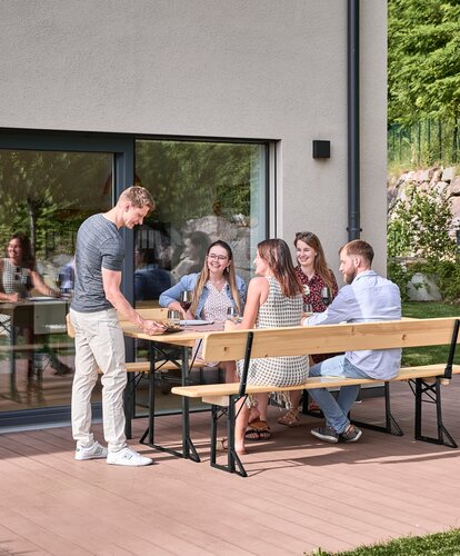 Freunde sitzen auf der breiten Bierzeltgarnitur mit Lehne in Natur und stoßen gemeinsam an.