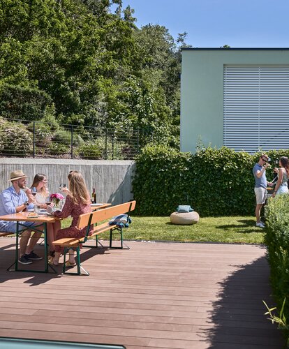 3 Personen sitzen auf einer breiten Bierzeltgarnitur mit Lehne in Kiefer im Garten und 2 andere Personen stehen neben dem Grill und unterhalten sich.