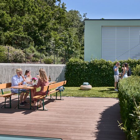 3 Personen sitzen auf einer breiten Bierzeltgarnitur mit Lehne in Kiefer im Garten und 2 andere Personen stehen neben dem Grill und unterhalten sich.