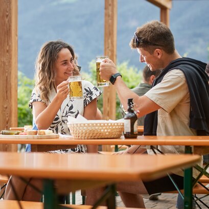 Ein Paar stoßt mit zwei Bierkrügen zusammen und sitzt dabei auf klassische Bierzeltgarnituren auf der Terrasse beim Guggerhof.