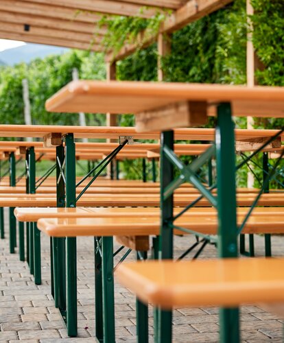 Reihe klassischer Bierzeltgarnituren mit grünem Untergestell und in Natur unter einer Pergola im Außenbereich, vor einer begrünten Steinmauer.