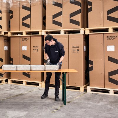 Eine Standaufnahme von einem Belastungstest eines klassischen Biertisches. Ein Mitarbeiter legt nacheinander Ziegel auf die Fläche des Tisches, um die Belastbarkeit zu prüfen.