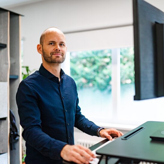 An employee of the Zingerle Group is standing at his ergonomic standing desk, looking into the camera. In front of him is his computer screen.