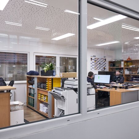Auf dem Foto sieht von außen in das Büro der Produktion hinein. Im Inneren sitzen drei Mitarbeiter an ihren Computern.