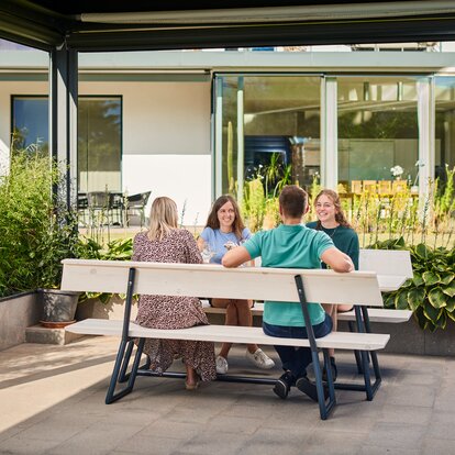 Man sieht eine Riva-Designgarnitur, die in einem Garten steht. Auf den beiden weißen Bänken mit Lehne sitzen vier Personen.