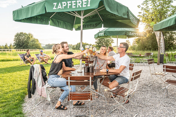 4 Personen stoßen mit ihrem Bier auf den Biergartenmöbeln auf der Terrasse eines Lokals an.