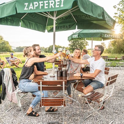 4 Personen stoßen mit ihrem Bier auf den Biergartenmöbeln auf der Terrasse eines Lokals an.