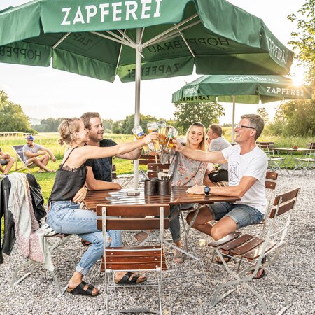 4 Personen stoßen mit ihrem Bier auf den Biergartenmöbeln auf der Terrasse eines Lokals an.
