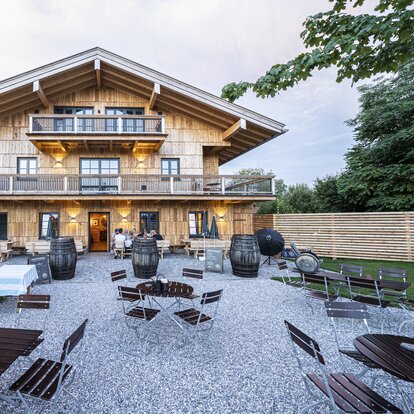 Auf dem Bild sieht man ein Gasthaus mit einer Holzfassade, davor ist ein großer Platz, wo eine Reihe von eckigen und runden Gartenmöbelgarnituren aufgestellt wurden.