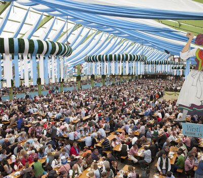 Auf dem Foto sieht man das Innere eines Oktoberfestzeltes. Tausenden Menschen sitzen auf RUKU1952® Bierzeltgarnituren. An der Decke befindet sich blau-weiße Deko.