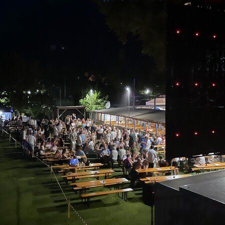 Das Bild zeigt das Public Viewing eines EM-Spiels bei Nacht. Viele Menschen sitzen auf Biergarnituren von RUKU1952® im Freien und schauen gemeinsam das Spiel. 