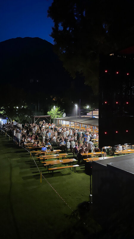 Das Bild zeigt das Public Viewing eines EM-Spiels bei Nacht. Viele Menschen sitzen auf Biergarnituren von RUKU1952® im Freien und schauen gemeinsam das Spiel. 